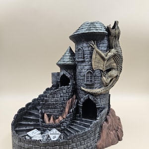 Pode incluir: Um tabuleiro de rolagem de dados de torre de dragão impresso em 3D com uma escada em espiral e um dragão esculpido no lado. A torre é feita de pedra cinza com detalhes em cobre. Existem cinco dados no tabuleiro.