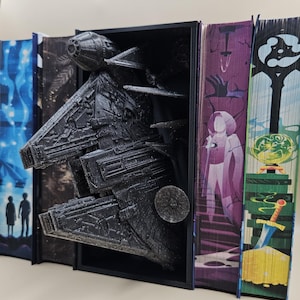 Può includere: Un modello di astronave stampato in 3D di colore nero è esposto in un ferma libri. L'astronave sporge parzialmente dal ferma libri. Il ferma libri è in legno e ha una cornice nera. L'astronave è dettagliata e ha una superficie testurizzata.