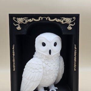 Op de afbeelding: Een wit uilenbeeldje in een zwart frame met gouden accenten. De uil zit op een zwarte basis en heeft een gedetailleerd, realistisch uiterlijk.