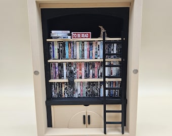 May include: A miniature wooden bookshelf diorama with a black interior. The shelves are filled with tiny books. A red sign reads "TO BE READ." A black ladder leans against the top shelf. A small cabinet with doors is at the bottom.