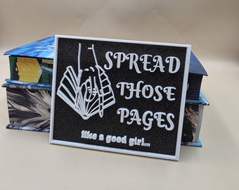 Op de afbeelding: Een zwart vierkant bord met witte tekst met de tekst "SPREAD THOSE PAGES" en "like a good girl...". Het bord heeft een witte rand en een witte illustratie van een boek. Het bord staat voor twee boeken.