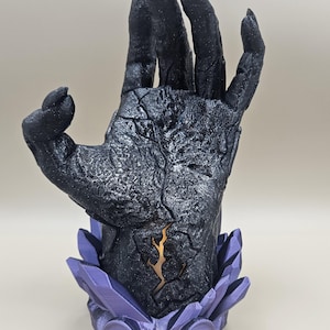以下が含まれることがあります： 黒色のテクスチャ加工された手の彫刻で、手のひらに金色のデザインが施されています。 手は幾何学模様の紫色の台座で支えられています。 彫刻は詳細でひび割れた外観をしています。