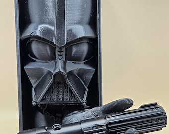May include: A black 3D-printed Darth Vader bust in a rectangular frame, with a black lightsaber held in a hand. The bust features the character's iconic helmet and mask. The lightsaber has a detailed handle.
