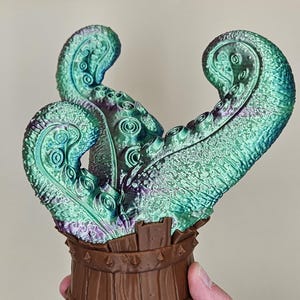 Könnte beinhalten: Eine dekorative Skulptur mit schillernden grünen und lila Tentakeln, die aus einem braunen Holzfass herausragen. Die Tentakel haben eine strukturierte Oberfläche mit kreisförmigen Mustern. Das Fass hat ein detailliertes Holzmaserungsdesign.