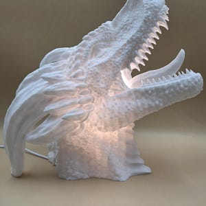 Pode incluir: Escultura de cabeça de dragão branca com a boca aberta, revelando dentes afiados e uma superfície texturizada. A escultura é iluminada por dentro, criando um brilho suave. Um cabo de alimentação estende-se da base. Lâmpada decorativa.