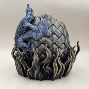 Peut inclure: Une figurine d'œuf de dragon noir et or avec un dragon qui émerge de l'œuf. L'œuf est texturé avec des écailles et le dragon a des ailes et une queue.