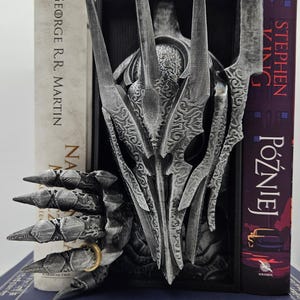 Può includere: Un ferma libri argentato e nero con un design dettagliato dell'Unico Anello de Il Signore degli Anelli. Il ferma libri tiene su due libri, uno intitolato "A Game of Thrones" di George R.R. Martin e l'altro intitolato "Stephen King - Później" in polacco.