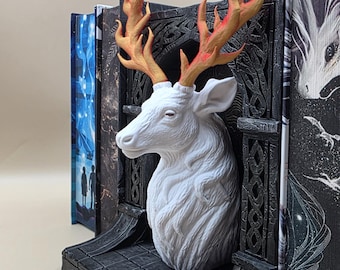 May include: A white deer head bookend with orange and gold antlers. The bookend is set against a dark gray, ornate background. Books with colorful covers are placed on either side of the bookend, creating a decorative display.