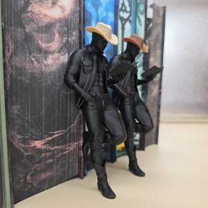 May include: Two black cowboy figurines, each wearing a hat and holding a notepad, lean against a stack of books. One cowboy wears a tan hat, the other a brown hat. The figurines are detailed with realistic clothing.