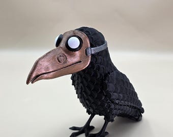May include: A black crow figurine with a copper-colored plague doctor mask and large white eyes. The crow has a textured body and stands on two black feet. The mask is secured with a silver band around the head.