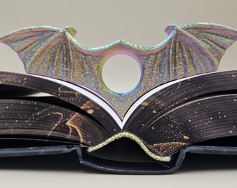 May include: A iridescent metal dragon wing bookmark with a hole in the center. The bookmark is resting on the spine of a closed book with a gold-colored edge.