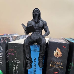 Puede incluir: Una figura negra impresa en 3D de una persona con una máscara de Scream leyendo un libro, posada sobre una pila de libros. La figura es musculosa y detallada, con una pose sentada. Los libros tienen cubiertas negras.
