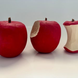 May include: Three wooden apple sculptures arranged in a row. The first is a solid red apple, the second is a red apple with a carved bite mark revealing light wood, and the third is a red apple core showing the light-colored interior wood.