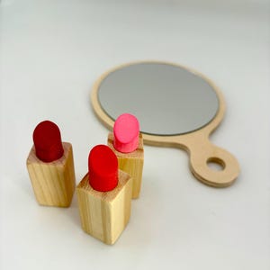 May include: Wooden toy set featuring three lipstick replicas with red and pink tops, alongside a small, round hand mirror. The lipsticks are set on small wooden blocks. The mirror has a wooden handle.