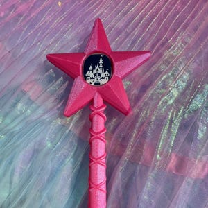 Könnte beinhalten: Rosaer Stern-förmiger Zauberstab mit einem Schwarzweißbild eines Schlosses in einem Kreis. Der Zauberstab hat einen rosa Griff mit einem Kreuzmuster und einen silbernen Schlüsselanhänger.
