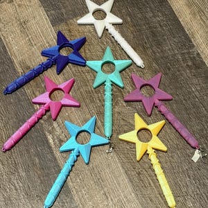 Könnte beinhalten: Eine Sammlung farbenfroher sternförmiger Zauberstäbe in verschiedenen Farben, darunter Weiß, Blau, Rosa, Grün, Lila, Hellblau und Gelb. Jeder Zauberstab hat einen strukturierten Griff und eine kreisförmige Öffnung im Stern.