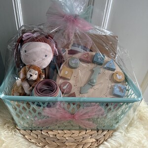 Könnte beinhalten: Ein Geschenk-Korb gefüllt mit einer Plüschpuppe, einem kleinen Löwen-Stofftier, einem Holz-Uhrenspielzeug und einem Stapel bunter Ringe. Der hellblaue Korb ist in Zellophan mit einer rosa Schleife verpackt. Der Korb steht auf einer gewebten Matte.