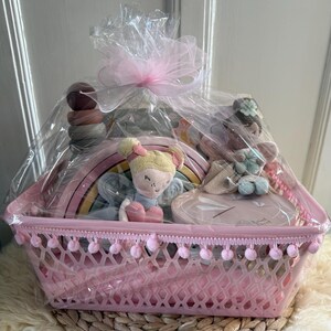 Könnte beinhalten: Ein rosa Geschenkkorb gefüllt mit Spielzeug, verpackt in durchsichtigem Zellophan und mit einer rosa Schleife gebunden. Der Korb enthält einen Holzregenbogen, ein Stapelspielzeug und zwei Plüschpuppen. Der Korb hat eine dekorative Bordüre mit rosa Pompons.