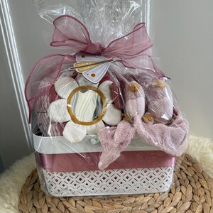 Könnte beinhalten: Ein Geschenk-Korb, verpackt in durchsichtigem Zellophan und mit einer rosa Schleife gebunden. Der Korb enthält ein Plüschtier, eine dekorative Blume mit Spiegelmitte und andere Artikel. Der Korb ist weiß mit einem rosa Streifen.