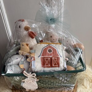 Könnte beinhalten: Ein Babygeschenkkorb gefüllt mit Plüschtieren, darunter ein Huhn, ein Lamm und ein weiches Buch in Scheunenform. Der Korb ist in durchsichtiges Cellophan gewickelt und mit einer blauen Schleife gebunden. Der Korb steht auf einer gewebten, kreisförmigen Basis.