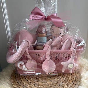 Könnte beinhalten: Ein rosa Babygeschenkkorb mit Spielzeug und Zubehör. Der Korb enthält eine Plüschpuppe, ein Holz-Steckspielzeug, eine Wal-förmige Rassel und ein sternförmiges Spiegelspielzeug. Der Korb ist in durchsichtiges Cellophan gewickelt und mit einer rosa Schleife gebunden.
