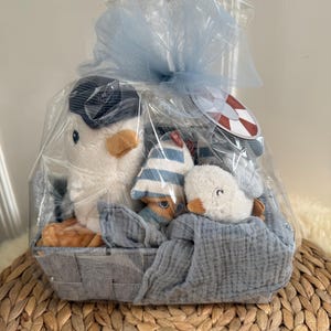 Könnte beinhalten: Ein Geschenk-Korb mit Plüsch-Pinguin-Spielzeugen, einem gestreiften Hut und einer blauen Decke. Der Korb ist in durchsichtiges Zellophan verpackt und mit einer hellblauen Schleife gebunden. Ein kleines Etikett mit einem Rettungsring-Design ist angebracht.