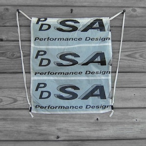 May include: A white drawstring bag with the text "PSA Performance Design" repeated three times in black. The bag is hanging from two white drawstrings attached to black metal hooks.