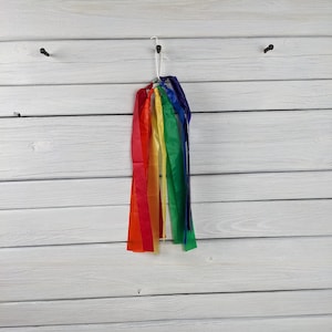 May include: A collection of colorful ribbons in a rainbow spectrum, including red, orange, yellow, green, and blue. The ribbons are hanging from a white hook against a white wooden plank background.