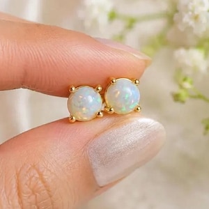Könnte beinhalten: Ein Paar runde Ohrstecker mit schillernden weißen Opal-Cabochons, die in polierten goldfarbenen Metallfassungen sitzen. Die runden Steine zeigen subtile grüne und blaue Lichtreflexe und werden von jeweils vier kleinen Metallkrallen gehalten.