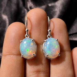 May include: A pair of drop earrings featuring oval-shaped, iridescent white opals set in silver-toned metal prongs. Each opal displays a play of color with flashes of green, orange, and blue. The earrings hang from simple silver-toned metal hooks.