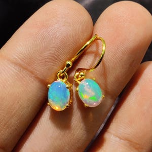 Può includere: Un paio di orecchini pendenti color oro. Ogni orecchino presenta una gemma di opale ovale con un gioco di colori, tra cui sfumature blu, verdi e gialle. Gli orecchini sono tenuti dalle dita di una persona.