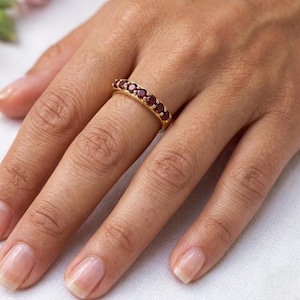 May include: A gold ring with a row of seven round, deep red gemstones. The ring is set on a finger, showcasing the jewelry's design and color. The ring's band is a warm gold tone, contrasting with the red stones.