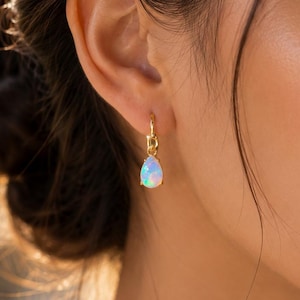 Peut inclure: Une boucle d'oreille créole en or avec un pendentif en opale en forme de goutte. L'opale présente des éclats iridescents de lumière bleue, verte et rose. La boucle est fixée par un petit anneau en or reliant l'anneau au sertissage en poire.
