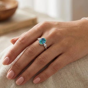 May include: A silver ring with an oval abalone shell center stone. The shell features swirling patterns of blue and green. The ring has decorative silver accents on the band. The ring is on a person's finger.