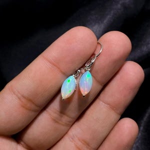 May include: A pair of silver drop earrings featuring oval-shaped opals. The opals have a white base with flashes of green and orange. The earrings are held by a person's fingers against a dark background.
