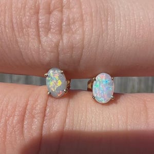 May include: A pair of oval-shaped opal stud earrings. The opals display a play of color with iridescent flashes of blue, green, and yellow. The earrings are set in gold-colored metal. The studs are held between fingers.