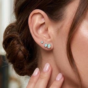 Puede incluir: Primer plano de la oreja de una mujer adornada con tres pendientes de ópalo. Los pendientes están engarzados en plata u oro blanco, con un pequeño perno redondo y dos ópalos ovalados más grandes. Los ópalos muestran un juego de colores.