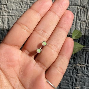 Puede incluir: Un par de pendientes de botón de ópalo redondos y pequeños. Los ópalos tienen un color base blanco con destellos verdes y amarillos. Los pendientes están engarzados en postes de plata.