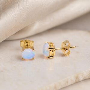 May include: A pair of gold stud earrings with oval-shaped opals. The opals have a milky white base with flashes of iridescent color. The earrings are set in a gold bezel setting with a post and butterfly backing.