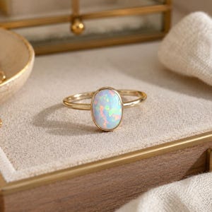 May include: A gold ring featuring a smooth, oval-shaped opal gemstone with iridescent flashes of blue, green, and pink. The stone is set in a simple gold bezel, and the band is a thin, polished gold wire, creating a clean and refined appearance.