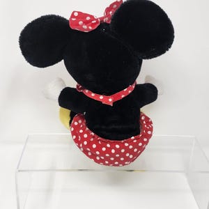 May include: Black plush Minnie Mouse toy with a red polka dot skirt and bow. The toy is sitting on a clear acrylic stand.