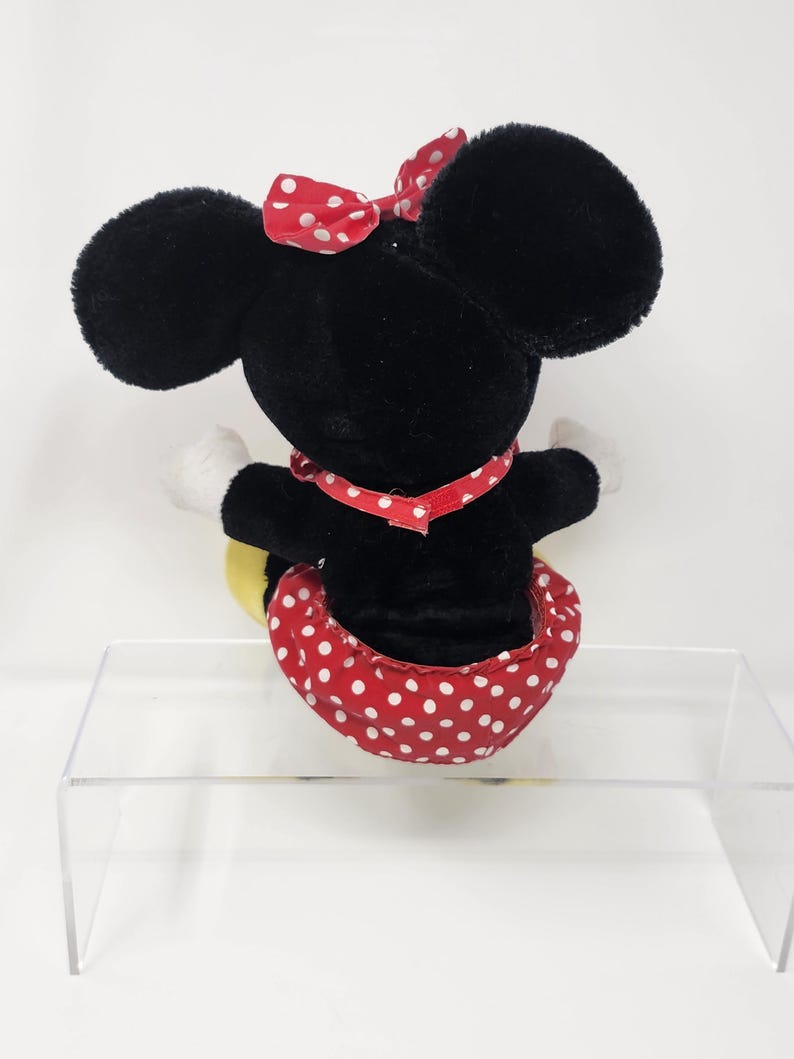 May include: Black plush Minnie Mouse toy with a red polka dot skirt and bow. The toy is sitting on a clear acrylic stand.