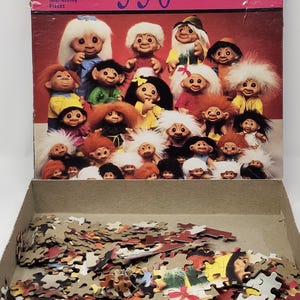 Puede incluir: Una caja de rompecabezas Golden Gallery con una imagen de Norfin Trolls. La caja está abierta, revelando piezas de rompecabezas. La caja dice "Family Reunion" y "550". Las piezas del rompecabezas están esparcidas en una bandeja de cartón.