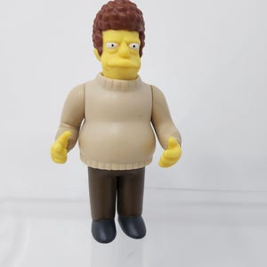 May include: A plastic action figure of a cartoon character with yellow skin, brown hair, and a beige sweater. The figure has brown pants and black shoes. The figure is standing upright with its arms at its sides. The figure is a collectible item.