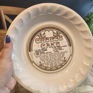 Peut inclure: Une assiette à tarte en céramique blanche avec un bord festonné, présentant une recette de cheesecake au centre. La recette est imprimée en marron et or. L'assiette est tenue par une personne.
