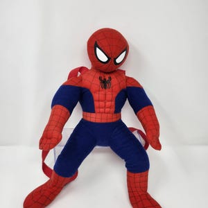 May include: A plush Spider-Man character backpack featuring a red and blue fabric body with a black spider web pattern, white eye patches, and a black spider emblem on the chest. The item includes red adjustable straps attached to the back.