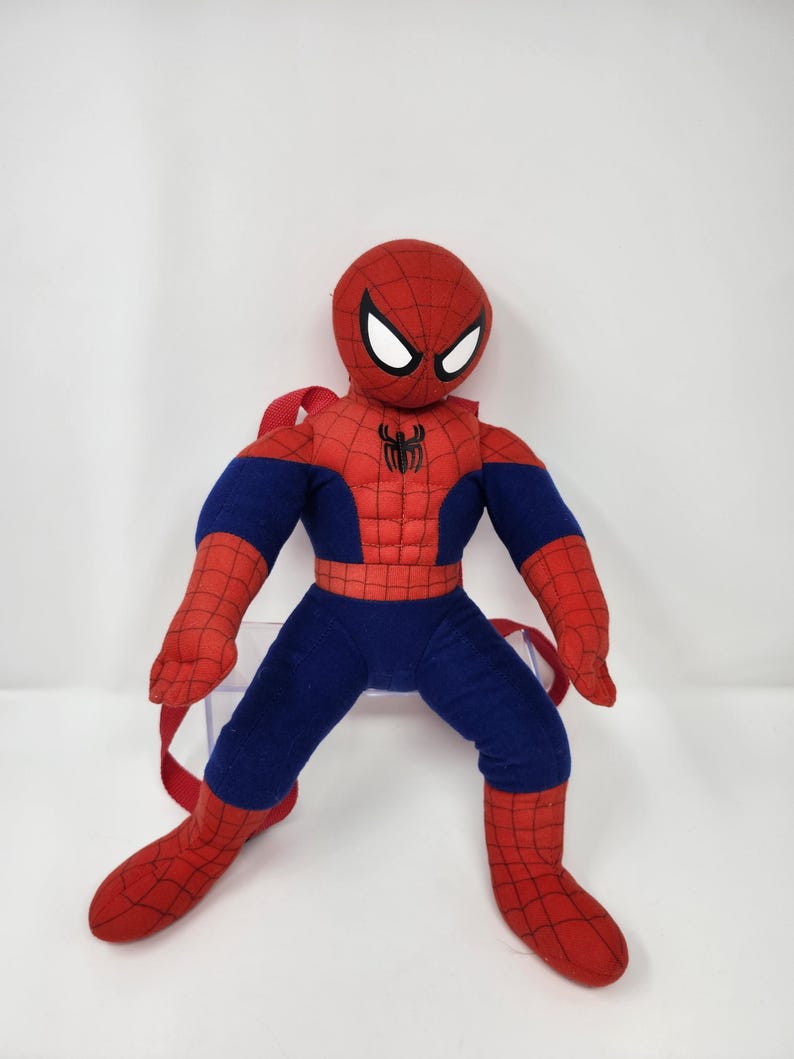 May include: A plush Spider-Man character backpack featuring a red and blue fabric body with a black spider web pattern, white eye patches, and a black spider emblem on the chest. The item includes red adjustable straps attached to the back.