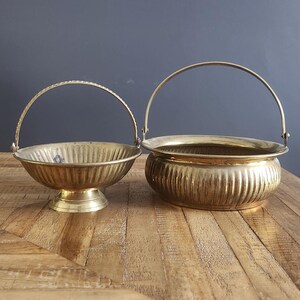 May include: Two brass bowls with handles. The bowls are on a wooden surface. The larger bowl is wider and has a more rounded shape. The smaller bowl is more narrow and has a more flared shape.