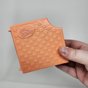 May include: An orange plastic Little Pet Shop accessory with a floral pattern and the brand name. The item has a rectangular shape with a curved edge and small protrusions. The surface is covered in embossed flower shapes.