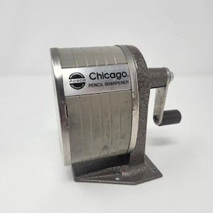 May include: A vintage Chicago Pencil Sharpener, made of silver and gray metal. The sharpener has a crank handle and a base for stability. The text "Chicago. PENCIL SHARPENER" is visible on the front.